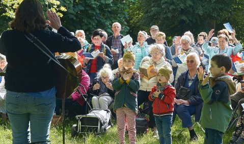 Viele Familien (kleinere Kinder in der ersten Reihe) draußen im Grünen. Ihnen zugewandt steht jemand mit Gitarre und ruft ihnen etwas zu.
