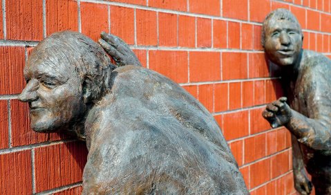Zwei Bronzen an einer geklinkerten Wand: Die Figuren (zwei ältere Männer) drücken ihre Ohren an die Wand.