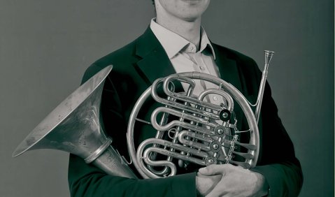 Grau-Stufen Foto eines jungen Erwachsenen Hornisten in Hemd und Sakko. Er trägt das Horn 103 von Alexander im Arm.