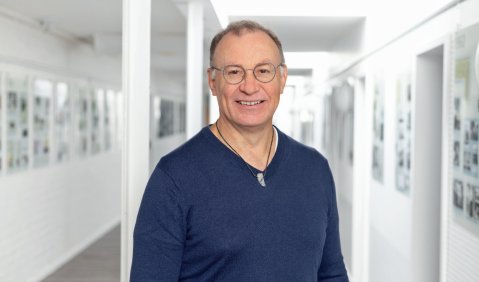 Ein Mann mit sehr kurzen Haaren und blauem Oberteil l&auml;chelt in einem wei&szlig;en Flur an dessen W&auml;nde einige Fotos h&auml;ngen.