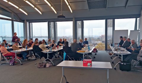 Ein offen gestalteter Tagungsraum mit zwei Fensterseiten in einem hohen Geschoss mit Blick über die Stadt. Darin 5 lange Tische mit DTKV Mitgliedern.