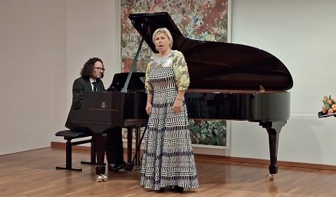 Eine Sängerin in einem stark gemusterten langen weiß-blau-grünen Kleid steht in der Ausbuchtung eines Flügels. Am Flügel ein Mann im dunklen Anzug mit langen dunklen Haaren.