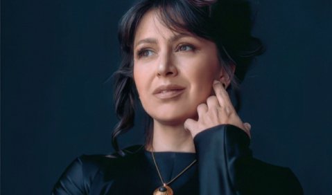 Eine Frau mit schwarzen Haaren in einem dunkelblau schimmernden Oberteil. Eine Hand hält sie seitlich an ihr Kinn und schaut in die andere Richtung.