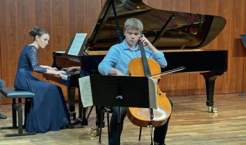 Ein Cellist (kurze Blond Haare, hellbraunes Cello, hellblaues Hemd) und eine Pianistin (dunkelblaues Kleid, Dutt).