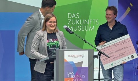Eine Frau in einem grauen Blazer an einem Rednerpult mit der Aufschrift "Das Zukunftsmuseum". Im Hintergrund h&auml;lt ein Mann einen gro&szlig;en Scheck.