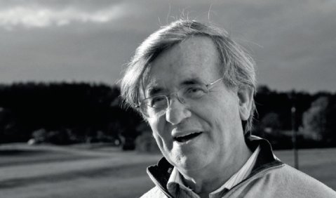 Schwarzwei&szlig;bild eines Mann im h&ouml;heren mittleren Alter mit Brille der auf einer leicht h&uuml;geligen Wiese freundlich in ein Gespr&auml;ch verwickelt ist.