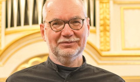 Ein Mann in schwarzem Hemd vor einer Orgel mit einem roten Buch mit einem goldenen handschriftlichen Schriftzug darauf.