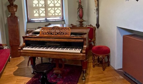 Ein historisches Zimmer mit großem Kruzifix und einem Portrait an der Wand. An der anderen Wand ist ein vergittertes Fenster. In der Ecke dieser beiden Wände steht der historische braune Flügel. Er sieht etwas kleiner aus als ein modernes Instrument.
