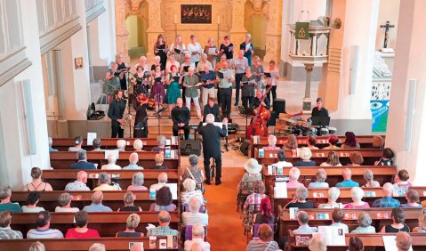 Ein gut gefülltes Kirchenschiff mit einem bunt gekleideten Chor im Altarraum. Vor dem Chor ist ein Ensemble platziert, dass an eine kleine Jazz-Combo erinnert.