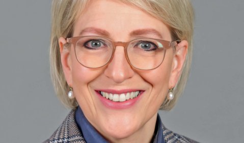 Ein hellblonde Frau mittleren Alters in kariertem Blazer, blaum Hemd, Brille und ernstem Lächeln.