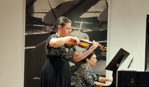 Eine Violinistin mit schwarzen Haaren in einem langen schwarzen Kleid steht leicht versetzt vor einer Pianistin. Beide auf einer leicht erhöhten hölzernen Bühne unter einer niedrigen Decke.
