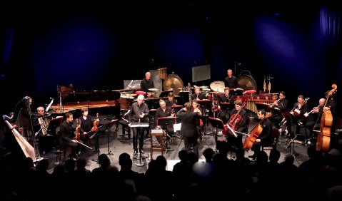 Ein Ensemble aus einer Vielzahl verschiedenster Instrumente (hauptsächlich Orchesterinstrumente), spielt hell erleuchtet in einem sonst sehr dunklen Raum.