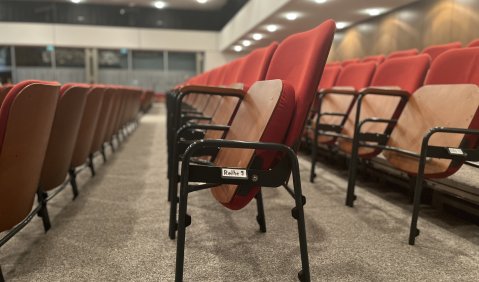 Orange-rot bespanntes, schlichtes Theatermobiliar. Decke und Wände wirken wie ältere Mehrzweckhalle.