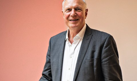 Ein Mann im grauen Anzug mit Halbglatze und dunkelgrauem Anzug steht an einem Geländer. Der Hintergrund ist Beige-Lachs-farben