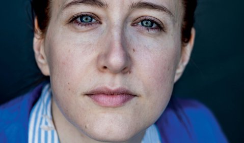 Porträt einer Frau die mit offenem und bestimmten Blick in die Kamera blickt. Sie ist in jungem mittleren Alter, trägt rote Haare, ein Hemd und einen blauen Pullover.