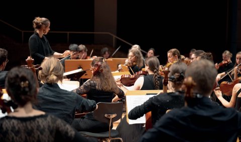 Foto der Streicherregister und Dirigentin aus der Perspektive der Kontrabässe also von rechts aus dem Orchester in das Orchester geguckt.