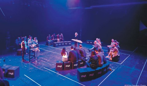Konzertperformance: Auf einer dunklen Bühne stehen sich zwei Musikerinnen an Schlaginstrumenten (Percussion und Hackbrett) gegenüber. Um sie herum in Halbkreisen sitzt das Publikum auf gepolsterten Hockern.