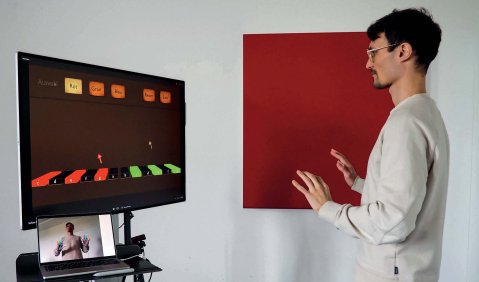 Ein junger Mann mit dunklen Haaren und kurzem Bart steht in hellem Pullover vor einem großen und einem kleinen Bildschirm. Der Große Bildfschirm zeigt mehrere farblich abgehobene längliche geometrische Figuren, an Klaviertasten erinnernd. Der aMann hält die Hände tastend vor sich.
