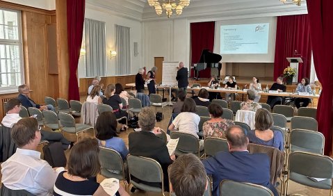 Eine Mitgliederversammlung in einem mittelgroßen und ehrwürdigen weiß gestrichenen Salon.