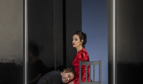 Tatjana im roten Kleid sitzt auf einem Stuhl. Eugen hockt vor ihr, den Kopf im Schoß. Um  beide herum ein Leuchtrahmen auf dem sonst dunklen Ausschnitt.