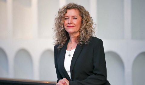 Eine Frau mittleren Alters mit blonden Locken in weißem Top und schwarzen Blazer lehnt an einem Geländer. Im Hintergrund eine weiße Wand aus Torbögen.