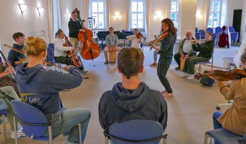 In einem schicken alten Schlosssalon sitzt ein Kreis verschiedener Menschen an verschienen Instrumenten zusammen oder steht.