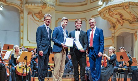 Die vier Herren unterschiedlichen Alters (zwei jüngere in der Mitte, zwei ältere außen) stehen in Anzügen oder vergleichbarem vor einem Orchester in einem barocken/klassizistischen Saal.