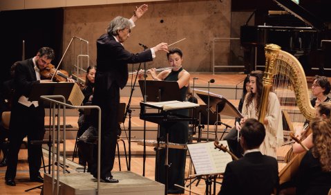 Dirigent mit erhobenen Armen vor einem Orchester, Musiker*innen spielen Instrumente wie Geige, Harfe und Fl&ouml;te. Foto: &copy; Berliner Festspiele, Fabian Schellhorn