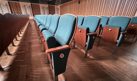 Theaterstuhlreihen aus Holz mit Tannengrün-blauen Polstern in einem rechtwinkligen Saal.