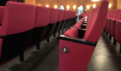 Rote Polster-Theaterstühle mit schwarzem Gestellt auf schwarzem Boden. Im Hintergrund eine cremefarbene Wand mit reichlich Leuchten. An der fotografierten Stuhlreihe eine Nummer: 9.