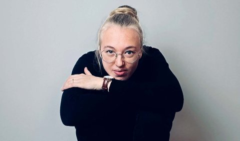 Eine junge blonde Frau mit Brille und schwarzen Klamotten sitzt vor einer weißen Wand auf einer Leiter. Die Arme auf den hohen Knien verschränkt abgestützt.