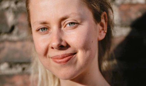 Eine unge Frau mit hellbraunen Haaren lächelt von der Sonne angeschien in die Kamera. Im Hintergrund eine Backsteinwand.