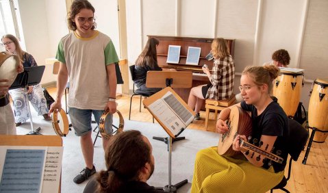 Mehrere junge Erwachsene machen erste Schritte auf verschiedensten Instrumenten. Eine Person geht herum und beobachtet die Fortschritte.