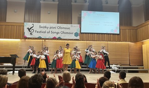 Ein Kinderchor in bunten Kleidern auf einer Bühne mit holzvertäfelter Rückwand.
