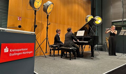 Ein Pianist und eine Querflötistin in schwarzer Abendkleidung auf einer Bühne mit Holzrückwand und Retro-Scheinwerfern.