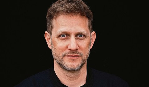 Ein Mann mit kurzen dunkelblonden Haaren und kurzem Bart in schwarzer Kleidung vor schwarzem Hintergrund.