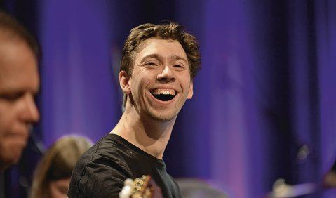 Schnappschuss eines strahlenden und musizierenden jungen Mannes. Welches Instrument er spielt, ist nicht sichtbar. Der Hintergrund ist lila beleuchtet. 
