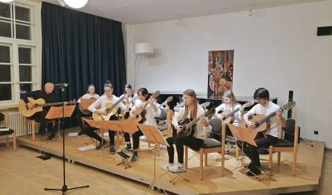 Eine große Anzahl junger Gitarristinnen und Gitarristen als Gitarrenorchester auf einer hölzernen Bühne in einem hellen Raum.