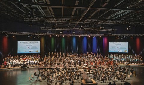 Musikschüler*innen und Schüler*innen aus Kooperationsschulen bilden gemeinsam mit den Musiker*innen der Hofer Symphoniker ein riesiges Orchester. Das Bild zeigt das Orchester in einer großen Veranstaltungshalle vor Publikum.