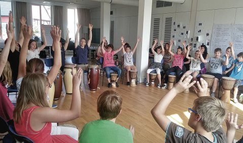 Ein Sitzkreis aus wenigen Erwachsenen und vielen Kindern mit Trommeln vor sich stehend. Alle haben die Hände nach oben gereckt.