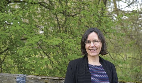 Lena Schwerdtner, eine Frau mit halblangem braunen Haar und Brille, steht lächelnd an einen Zaun gelehnt vor einem grünen Blättermeer.