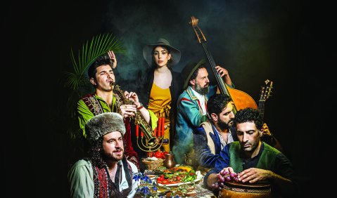 Eine bunt gekleidete Gruppe von rein schwarzhaarigen Menschen sitzen an einem gedeckten Tisch zusammen. Sie haben verschiedene Instrumente, von Klarinette, Trommel, Kontrabass, Gitarre und ein Keyboard beisammen. Manche der Instrumente sind zwischen dem Essen kaum zu erkennen.