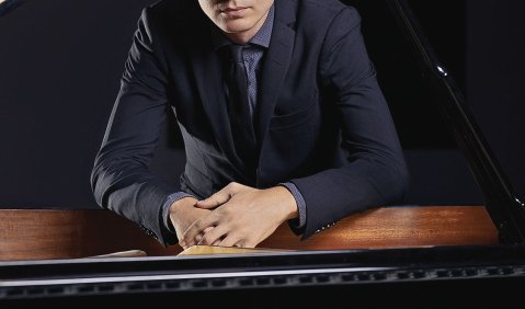 Der junge Pianist mit kurzem blonden Haar (leicht stehendem Pony) lehnt sich zum Portrait in den Flügel.