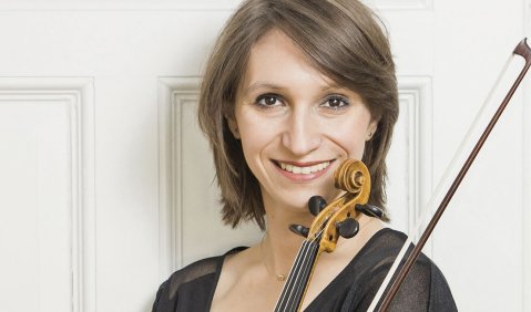 Eine Violinistin, ihr Instrument und Bogen in den Armen, steht in typischer Konzertkleidung mit dunklen braunen fast Schulterlangen Haaren vor einer weißen Tür.