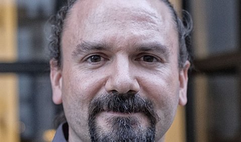 Ein Mann mit langen zum Pferdeschwanz gebundenen schwarzen Haaren und einem Bart um den Mund.