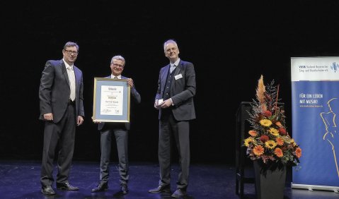 Ein Herr hält die Urkunde, flankiert von zwei weiteren Herren.