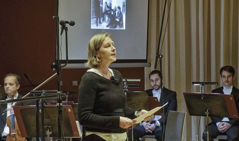 Im Hintergrund ein Kammerensemble und eine Leinwand mit einem angeworfenene Foto. Vorne Spricht eine Frau.