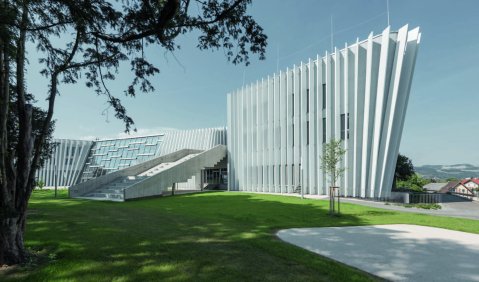Die Gebäude Anton Brucker Privatuniversität: Weiße geometrische Formen aus vielen Planken. Hauptaugenmerk ist ein Gebäude wie ein schmaler Keil mit Rippen. Eine Lange flache Treppe führt direkt ins erste Obergeschoss. Davor grüne Wiese im Hintergrund etwas tiefer liegend die Stadt.