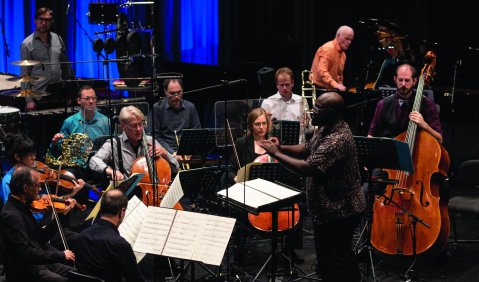 Vimbayi Kaziboni (ein Dirigent mit schwarzer Hauptfarbe) dirigiert ein Kammerorchester aus weißen Musiker*innen