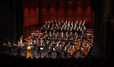 Auf einer großen zum Konzertsaal eingerichteten Theaterbühne tummeln sich die zahlreichen Musikerinnen und Musiker für die konzertante Aufführung: Orchester und Chor in schwarzer Kleidung mit weißen Hemden, die Solisten in ebenfalls schwarzen Anzügen, die Solistin in einem gelben Kleid.